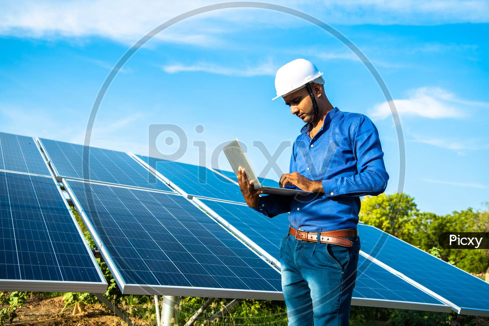 Young Male Engineer Working On Laptop Standing Near Solar Panels, Agriculture Farm Land With Clear Blue Sky Background, Renewable Energy, Clean Energy.