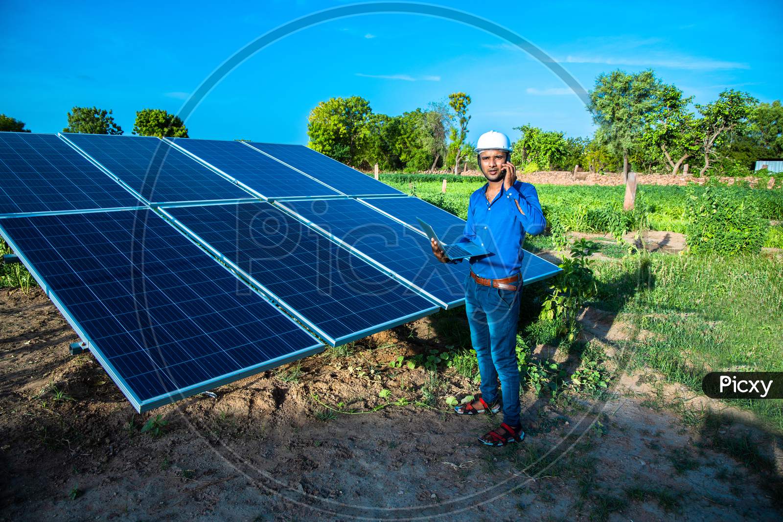 Young Male Engineer Talking On Phone With Laptop In Hand Standing Near Solar Panels, Agriculture Farm Land With Clear Blue Sky Background, Renewable Energy, Clean Energy.