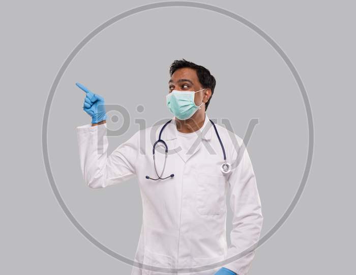 Indian Man Doctor Pointing To Side And Watching To Side Wearing Medical Mask And Gloves. Isolated