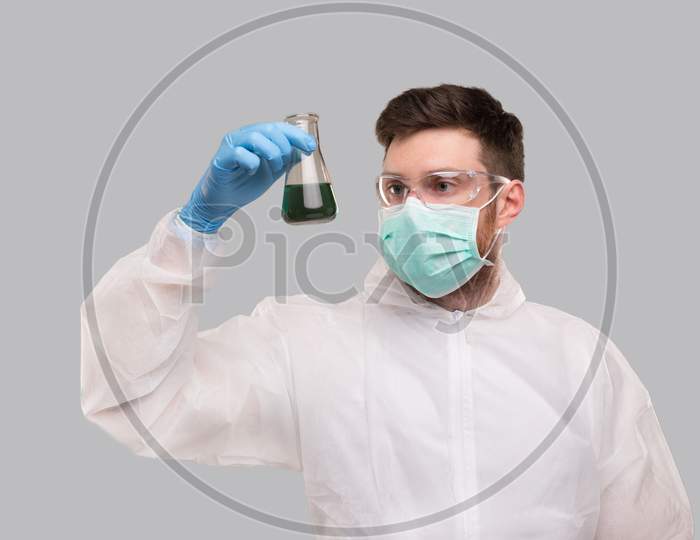 Male Laboratory Worker In Chemical Suit, Wearing Medical Mask And Glasses Watching Flask With Colorfull Liquid. Science, Medical, Virus Concept