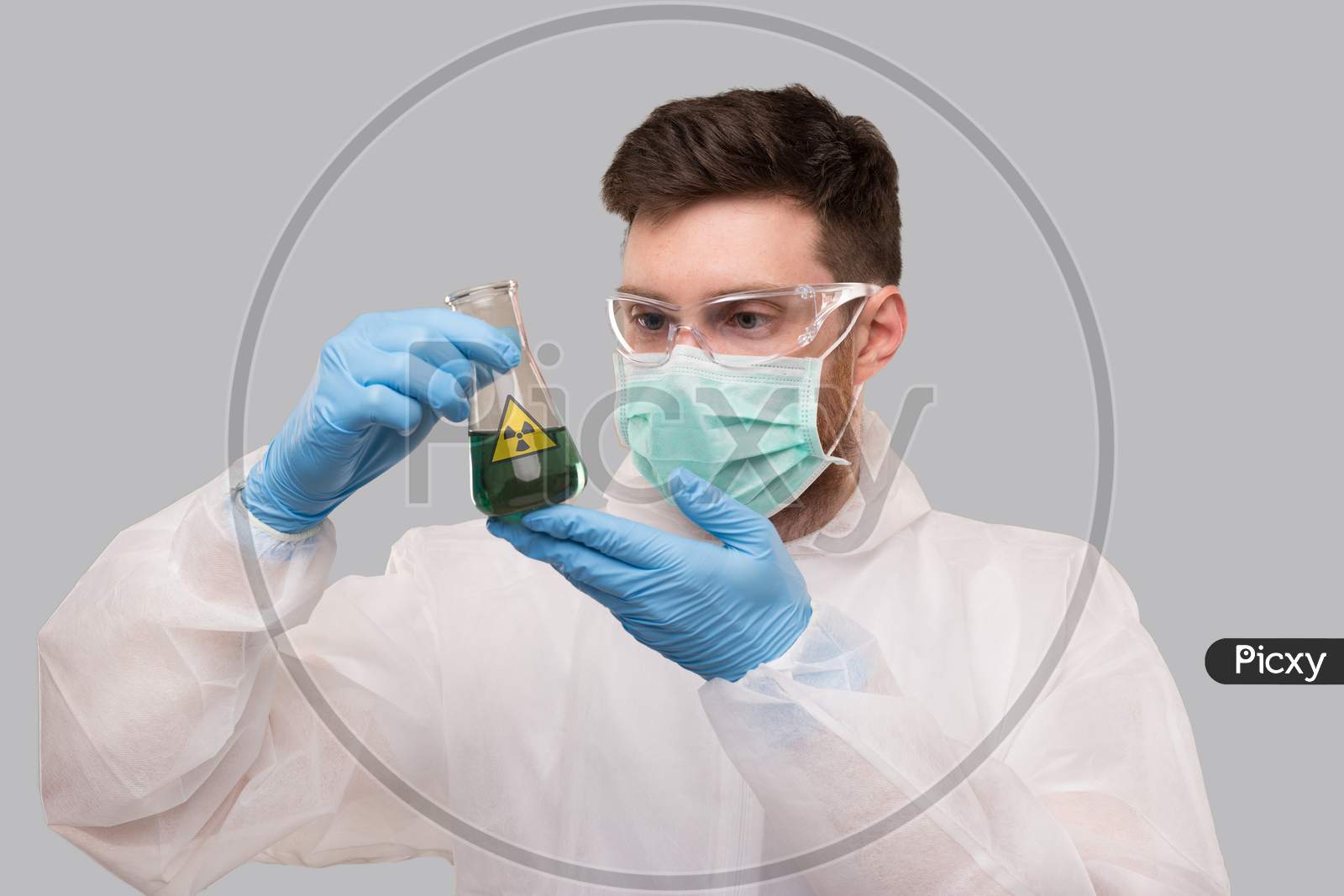 Male Laboratory Worker In Chemical Suit, Wearing Medical Mask And Glasses Watching Flask With Green Liquid Biohazard Sign. Science, Medical, Virus Concept