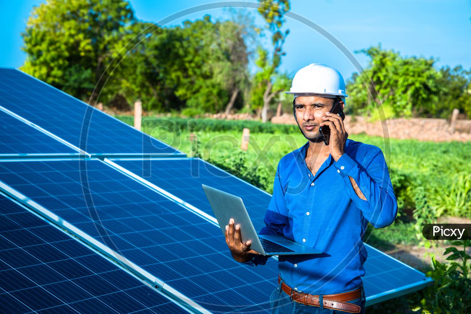 Young Male Engineer Talking On Phone With Laptop In Hand Standing Near Solar Panels, Agriculture Farm Land With Clear Blue Sky Background, Renewable Energy, Clean Energy.
