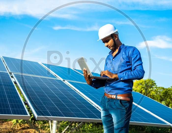 Young Male Engineer Working On Laptop Standing Near Solar Panels, Agriculture Farm Land With Clear Blue Sky Background, Renewable Energy, Clean Energy.