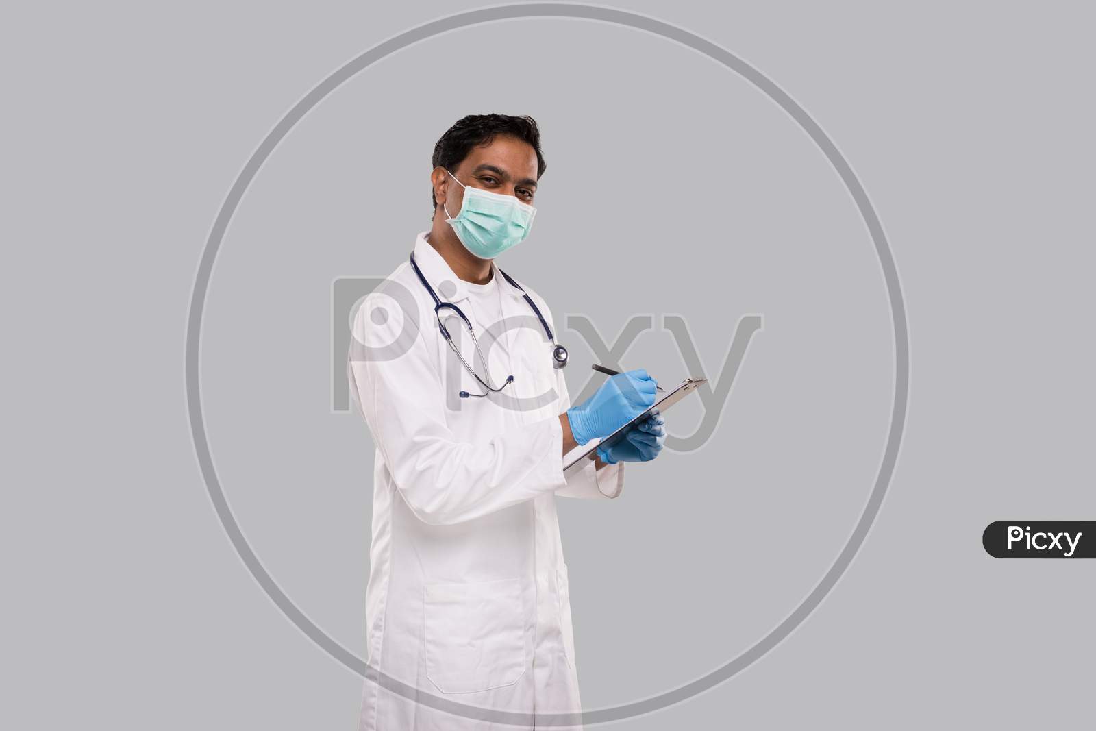 Doctor Writing In Clipboard Watching In Camera Wearing Medical Mask And Gloves. Indian Man Doctor Clipboard Isolated