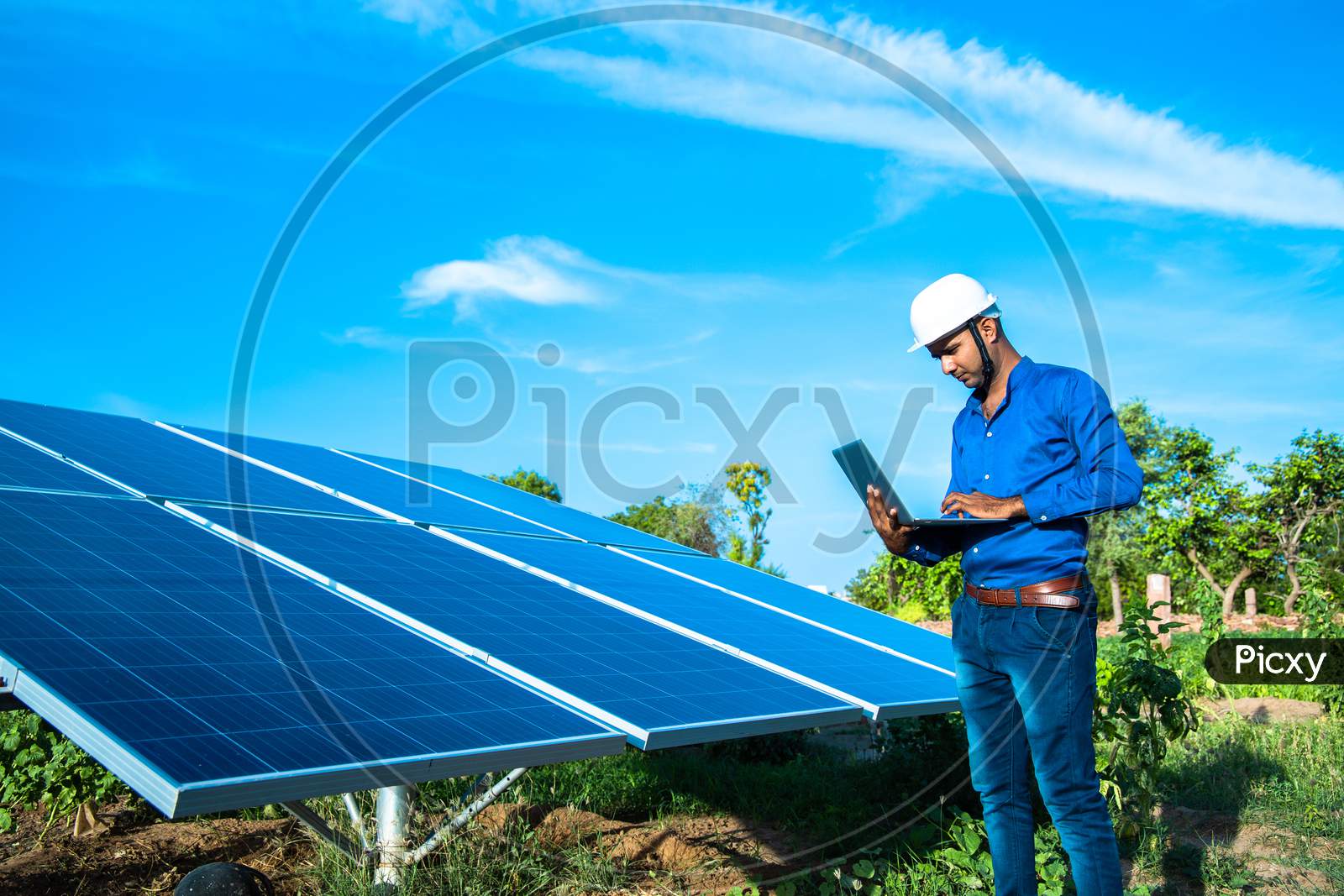 Young Male Engineer With Laptop In Hand Standing Near Solar Panels, Agriculture Farm Land With Clear Blue Sky Background, Renewable Energy, Clean Energy.