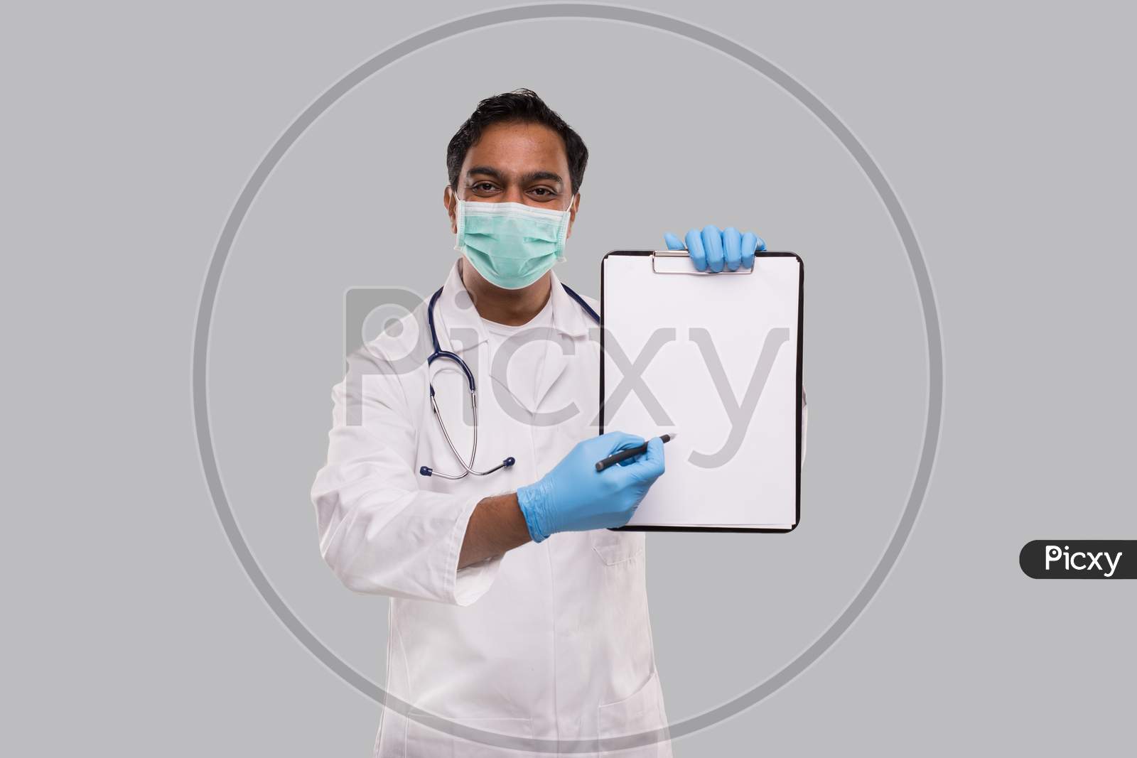 Doctor Pointing At Clipboard With Pen Wearing Medical Mask And Gloves Isolated. Indian Man Doctor Blank Clipboard In Hands