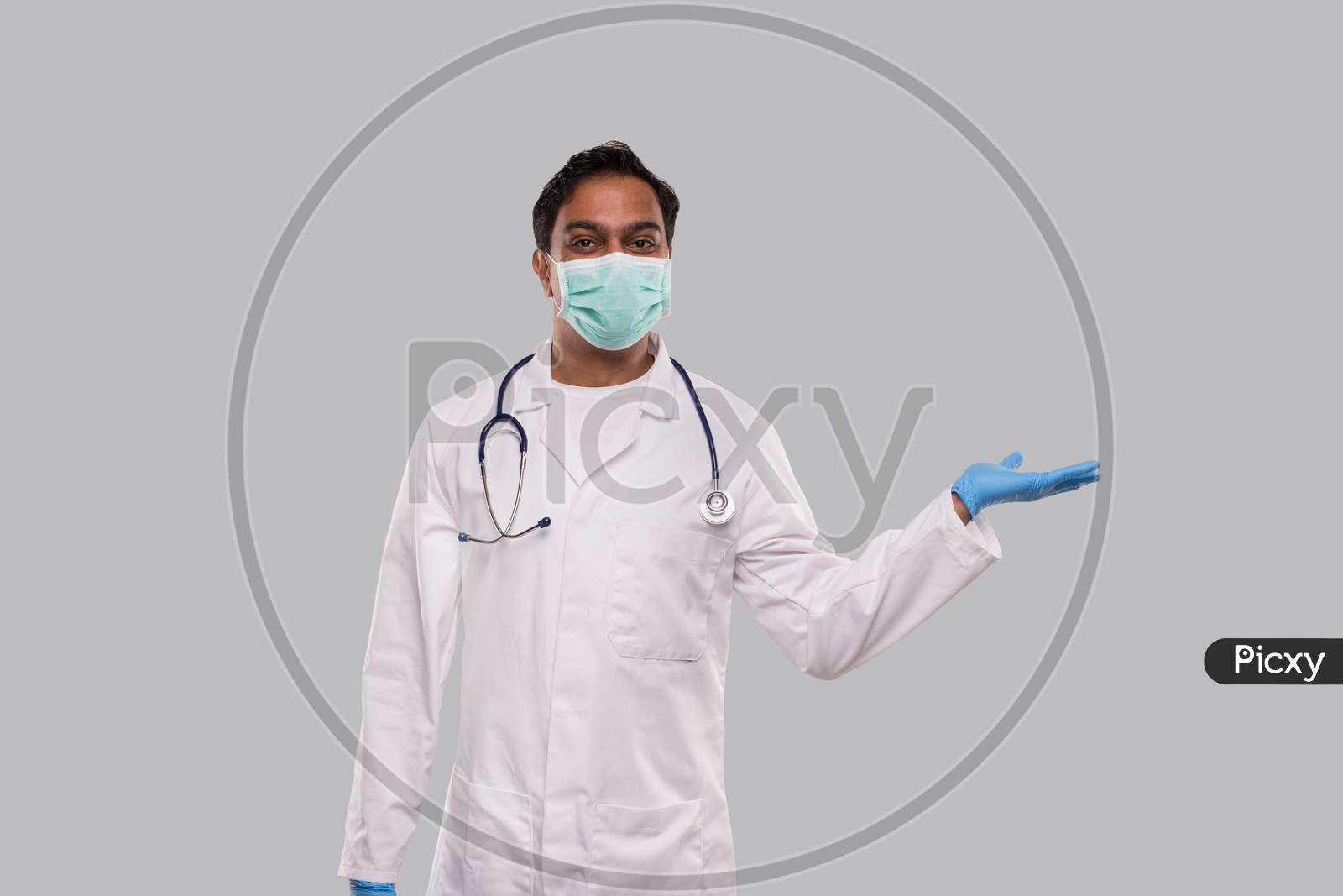 Doctor Holding Hand To Side Wearing Medical Mask And Gloves Watching To Camera Isolated. Indian Man Doctor Sign