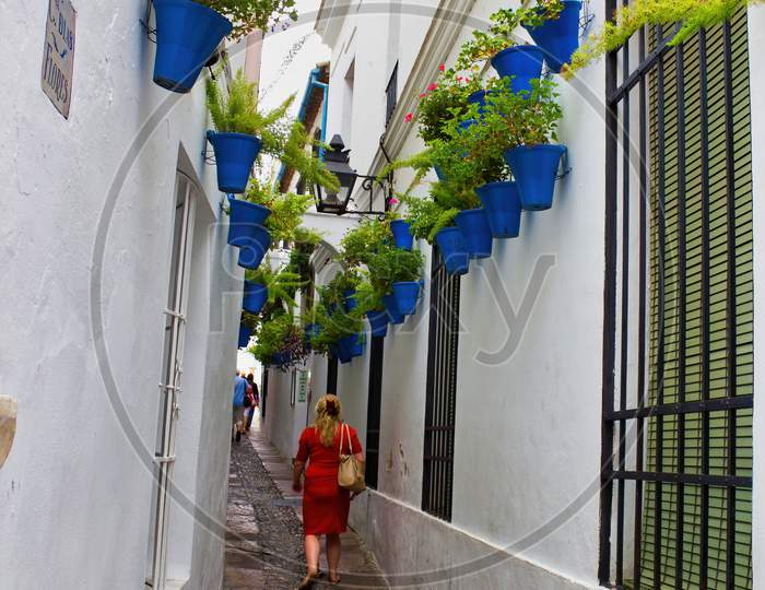 Cordoba, Spain - September 02, 2015: A Woman In Red Dress Walking Through Narrow Street With Flower Pots During Patio Festival Hanging On The White Wall In City Center Main Square,This Is One Of The Main Touristic Attraction