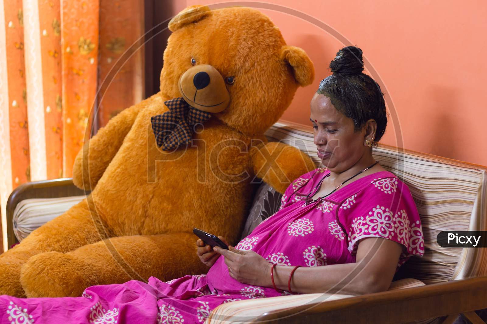 Senior Lady Has Applied Henna On Her Hair For Self Care. She Is Smiling And Watching Her Mobile Phone While Sitting On A Sofa Furniture Next To A Huge Orange Teddy Bear Indoors In Isolation.