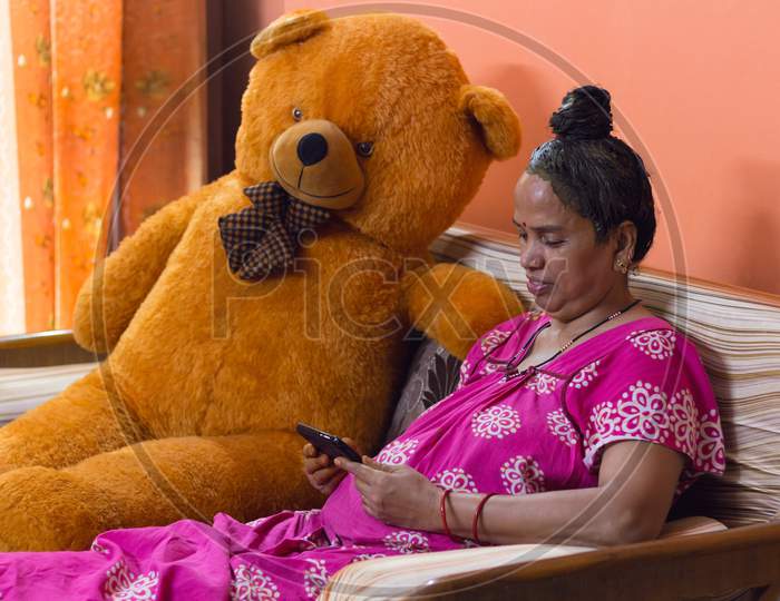 Senior Lady Has Applied Henna On Her Hair For Self Care. She Is Smiling And Watching Her Mobile Phone While Sitting On A Sofa Furniture Next To A Huge Orange Teddy Bear Indoors In Isolation.