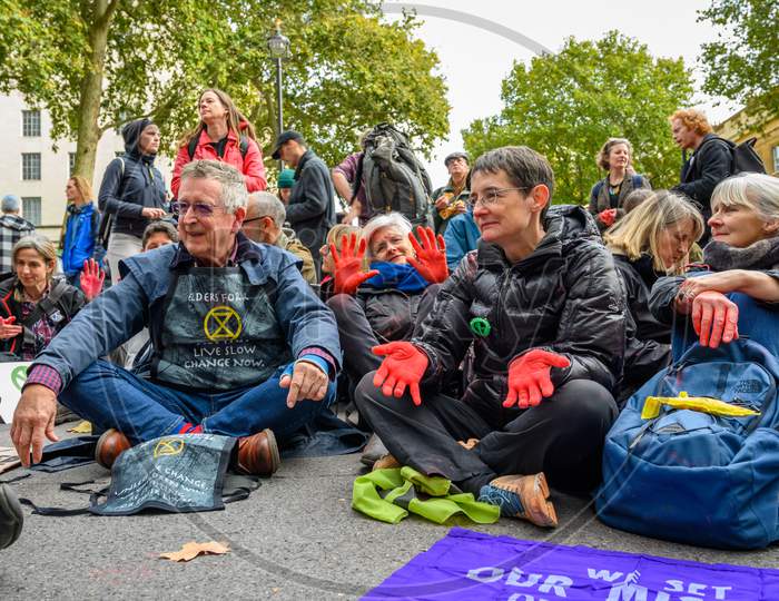 Mature Extinction Rebellion Protesters With Red Painted Hands Sat On A Road
