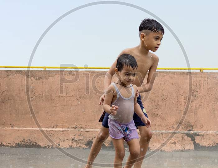 Two Cute Kids Wet, Playing And Having Fun In Rain At Their Terrace, Outdoors During Monsoon Season.