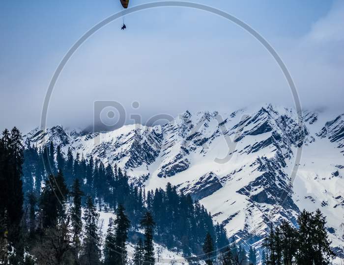 A Landscape Involving Snow Capped Valley With Pine Trees And Mountains. Paragliding Going On