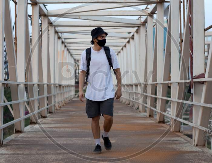 Young Millennial Walking On An Empty Metal Foot Bridge Outdoors While Wearing A Black Protective Face Mask To Prevent Covid-19 Virus Infection In A City. The New Normal.