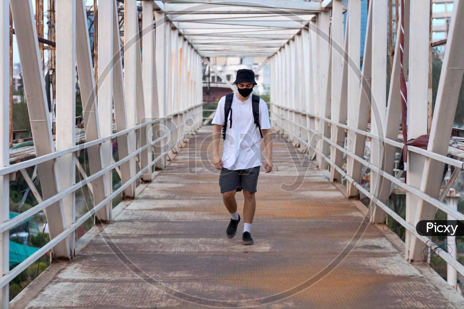 Young Millennial Walking On An Empty Metal Foot Bridge Outdoors While Wearing A Black Protective Face Mask To Prevent Coronavirus Infection In A City. The New Normal.