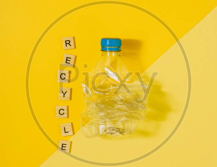 Transparent And Crushed Plastic Bottle With Blue Cap On A Yellow Background And The Word Recycle. Recycling And Environment Concept.