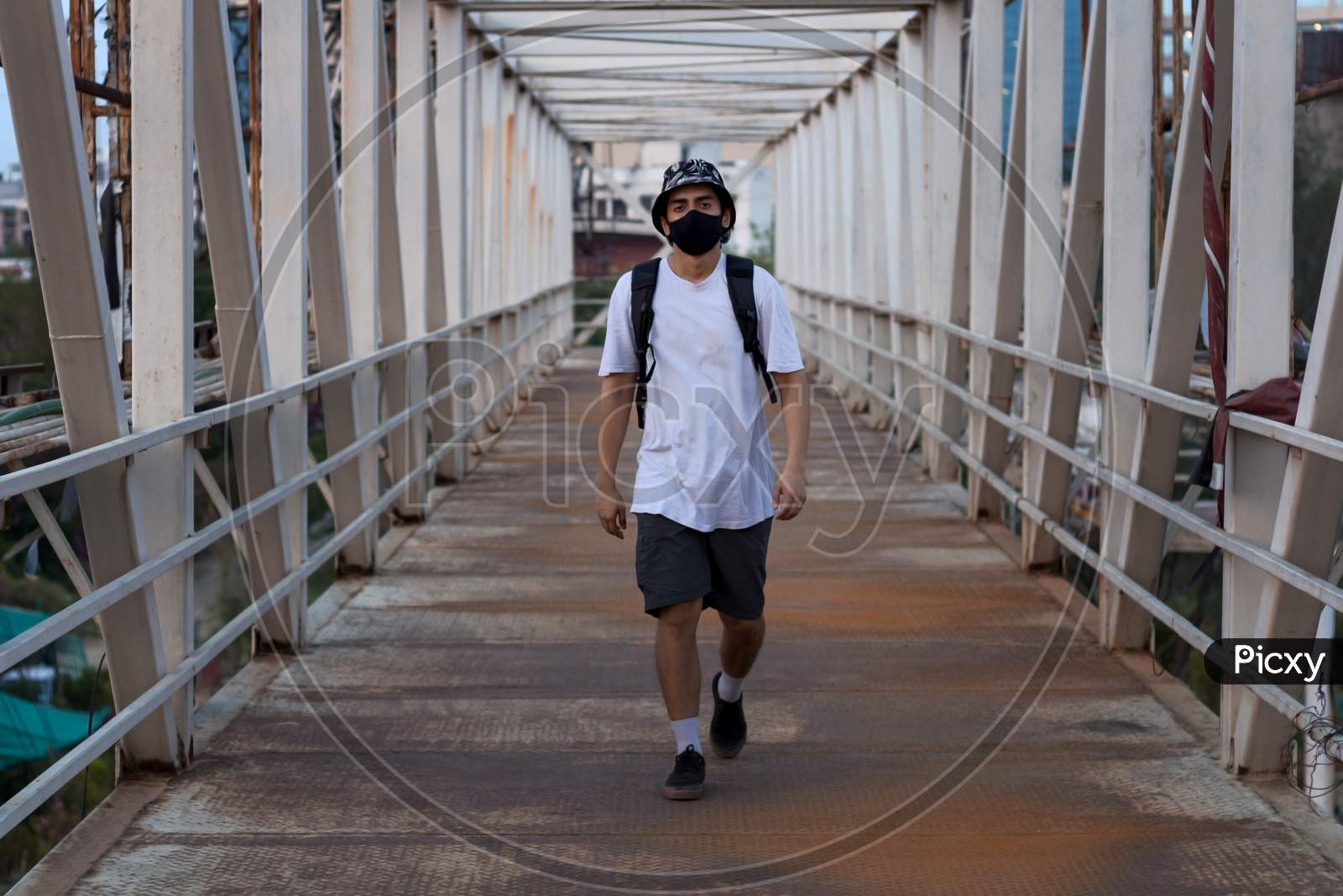 Young Millennial Walking On An Empty Metal Foot Bridge Outdoors While Wearing A Black Protective Face Mask To Prevent Covid-19 Virus Infection In A City. The New Normal.