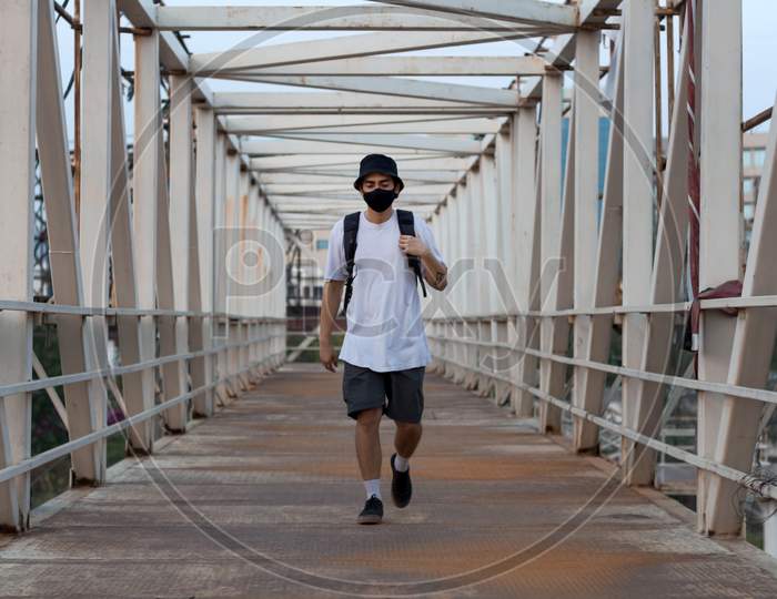 Young Millennial Walking On An Empty Metal Foot Bridge Outdoors While Wearing A Black Protective Face Mask To Prevent Corona Virus Infection In A City. The New Normal During World Pandemic.