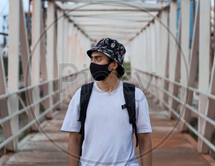 Portrait Of A Young Millennial On An Empty Metal Foot Bridge Outdoors While Wearing A Black Protective Face Mask To Prevent Covid-19 Virus Infection In A City. The New Normal.