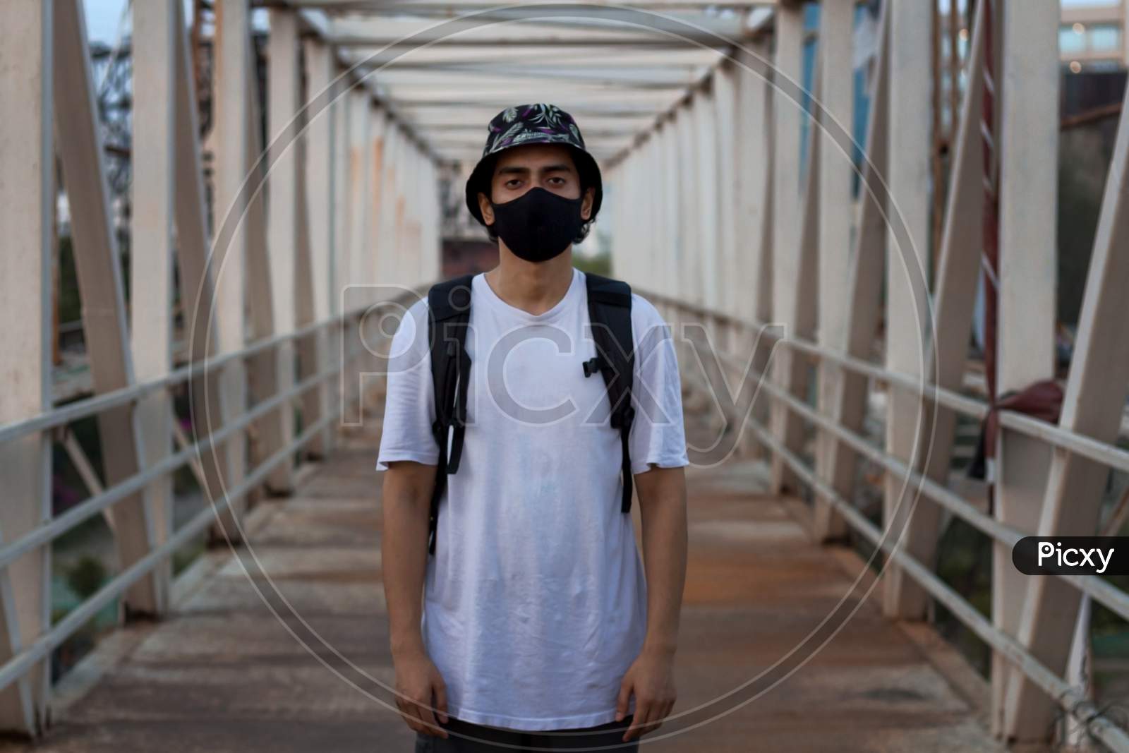 Young Millennial Standing On An Empty Metal Foot Bridge Outdoors While Wearing A Black Protective Face Mask To Prevent Coronavirus Infection In A City. The New Normal.