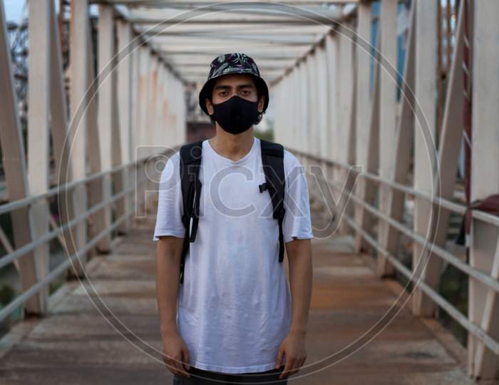 Young Millennial Standing On An Empty Metal Foot Bridge Outdoors While Wearing A Black Protective Face Mask To Prevent Coronavirus Infection In A City. The New Normal.