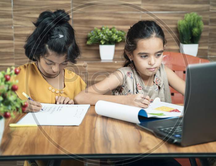 Two Kids Busy In Writing By Looking Into The Laptop During Online Or Virtual Class At Home - Concept Of E-Learning Or Distance Learning Or Children Lifestyle During Coronavirus Or Covid-19 Pandemic