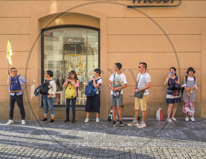 Asian Tourists Lined Up Behind A Flag Carrying Tour Guide On The Cobbled Streets Of The Old Town Quarter Of Prague, Czech Republic
