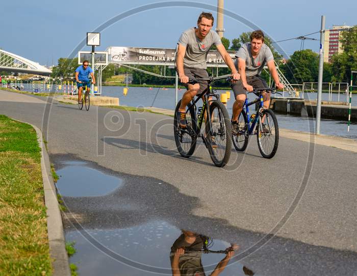 Cyclists On A Riverside Path With Reflection In Water Puddle