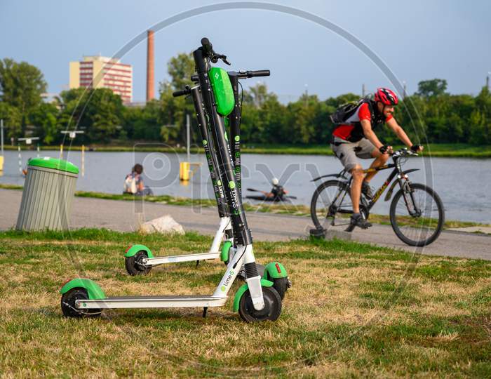 Electric Rental Scooters Parked Next To A Trash Can At The Side Of A River With Cyclist Passing In The Background