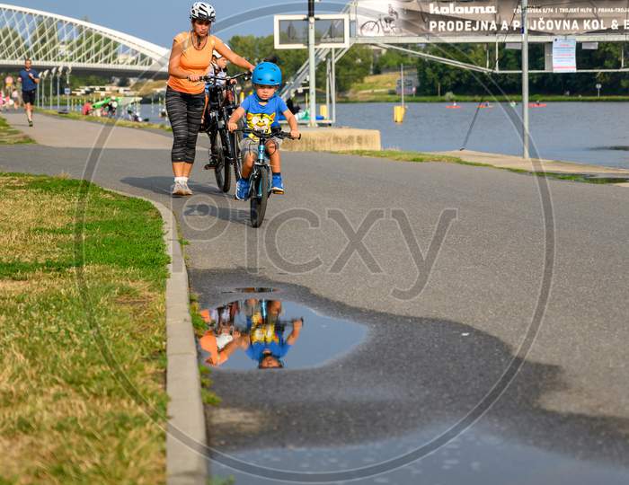 Young Child On A Bike Reflected In A Puddle Of Water And Followed By His Mother Pushing A Bicycle