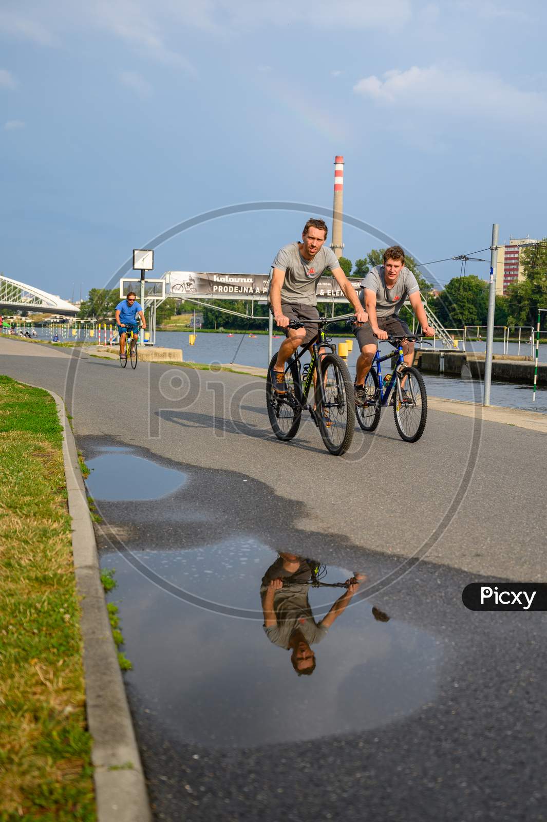 Cyclists On A Riverside Path With Reflection In Water Puddle