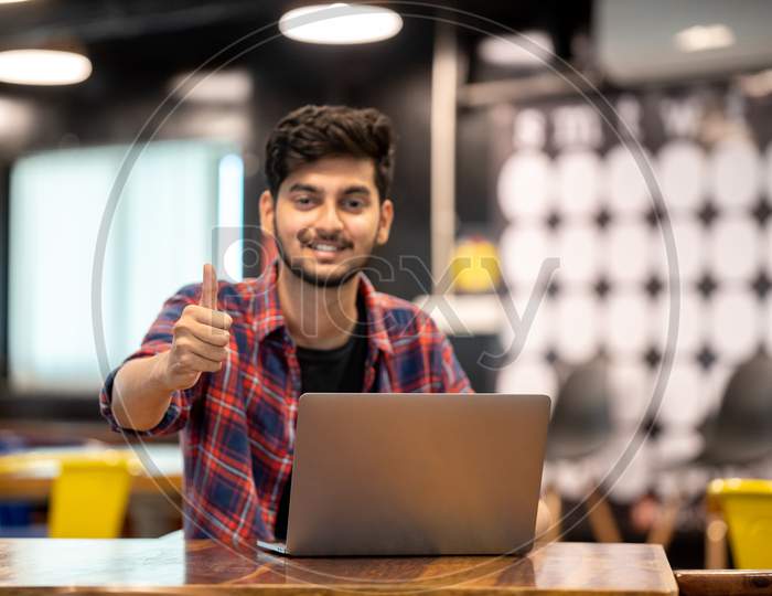 A Young Indian Man showing 'thumbs up' gesture while working on a laptop.