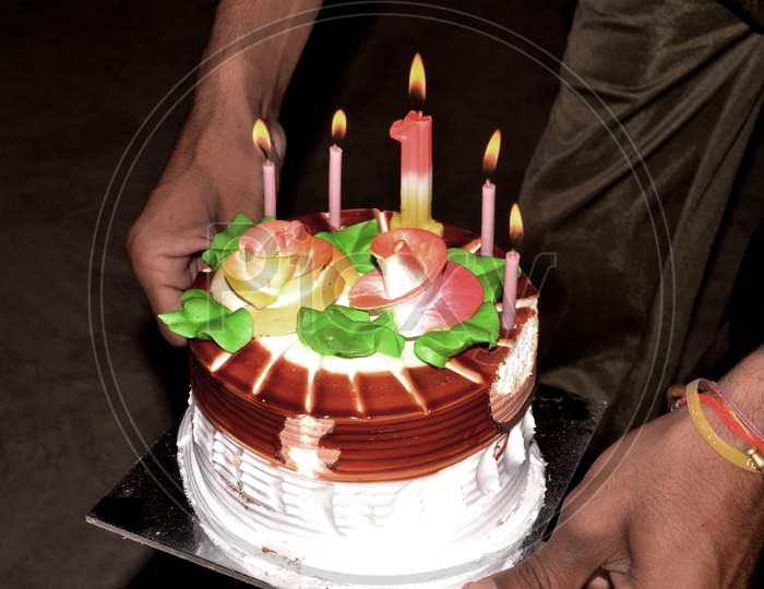 Birthday Drip Cake Holding In Hands And Burning Candle With Chocolate And Sprinkles Isolated Black Background Banner With Party Decor. Celebration Concept. Trendy Drip Cake