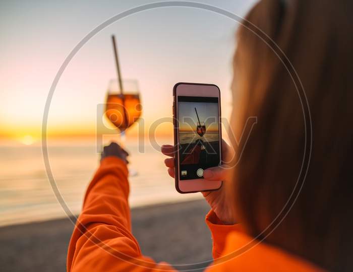 Girl Holding Glass Of Drink On Beach At Beautyfull Romantic Sunset Taking Pictures With Phone. Glass And Hand Close Up. View From Back