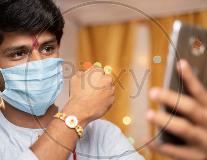 Man In Medical Mask Busy On Mobile Phone And Showing Rakhi Or Rakshabandhan To Sister Or Family Friends At Festival Ceremony During Coronavirus Or Covid-19 Pandemic At Home With Decoration Lights.