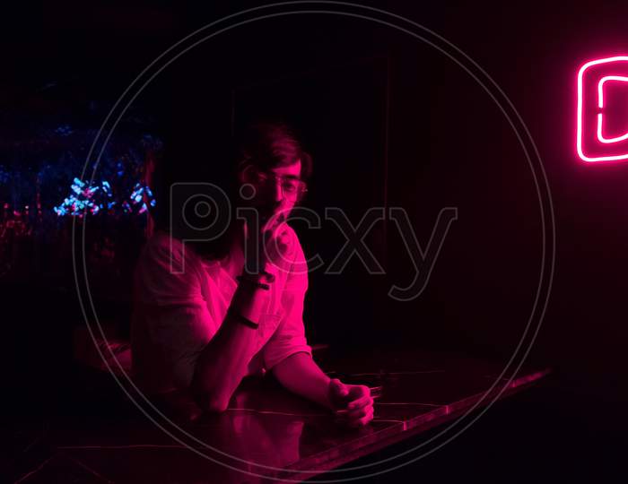 A Young Drunk Hipster Looking Man Looking Straight While Sitting Inside A Club Under A Bright Glowing Purple Neon Light.