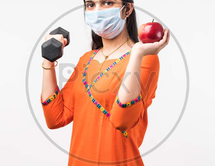 Indian Pretty Girl Exercising With Dumbbell And Showing Fresh Apple While Wearing Medical Face Mask