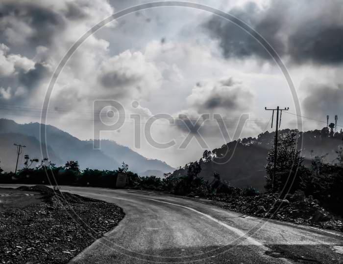 A monochrome picture of an empty road taking turn with thick clouds in the sky