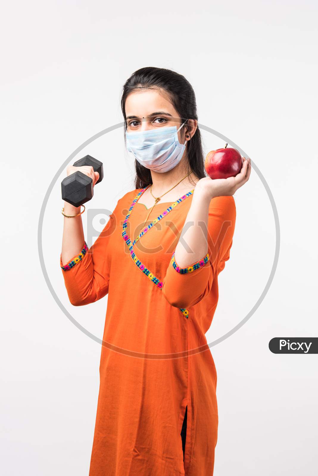 Indian Pretty Girl Exercising With Dumbbell And Showing Fresh Apple While Wearing Medical Face Mask