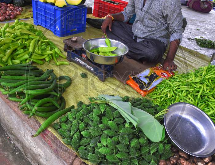 A Senior Indian Citizen Trades Vegetables In The Market. A Man Weighs Green Chili