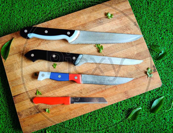 Four Colored Different Sizes Of Knives Kept On Chopping Board. With Curry Leaves And Coriander Leaves Spread Around