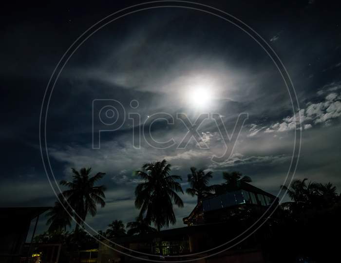 A Full Moon Night In A Village With Clouds Above And Buildings And Trees Below With Halo Surrounding The Moon