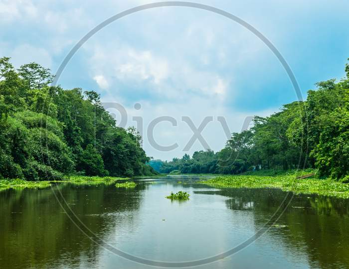 A Landscape Of A River With Transparent Water Flowing Through A Village With Surrounding Greenery. Blue Sky Above With Its Reflection On The Water