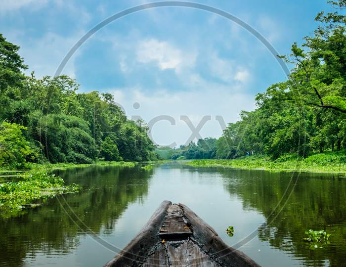 A Landscape Of A River With Transparent Water Flowing Through A Village With Surrounding Greenery Captured From A Boat. Blue Sky Above With Its Reflection On The Water