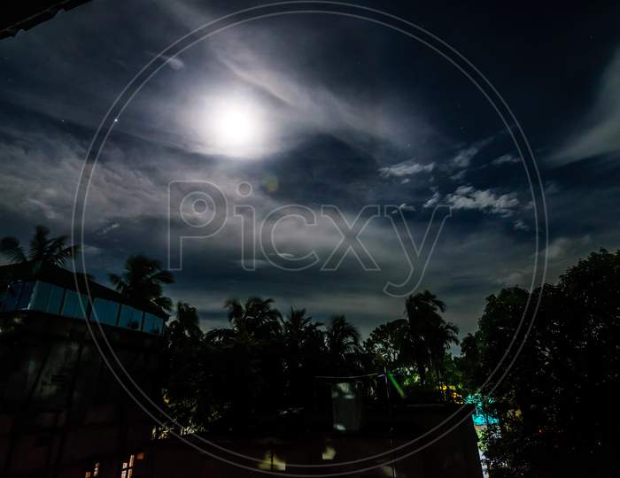 A Full Moon Night In A Village With Clouds Above And Buildings And Trees Below With Lights Peeping Through The Trees