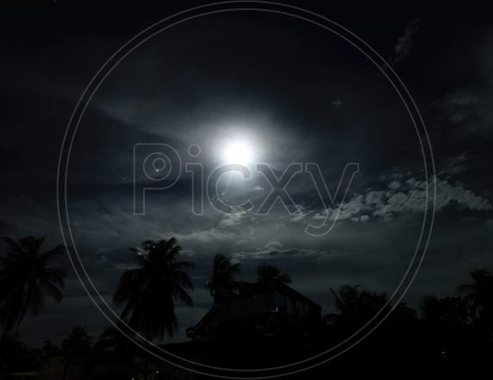 A Full Moon Night In A Village With Clouds Above And Buildings And Trees Below With Halo Surrounding The Moon
