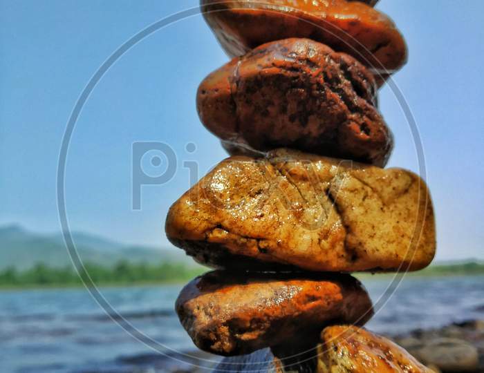 Stone Balancing Of Different Sizes Near A River With Water Flowing And Mountains.