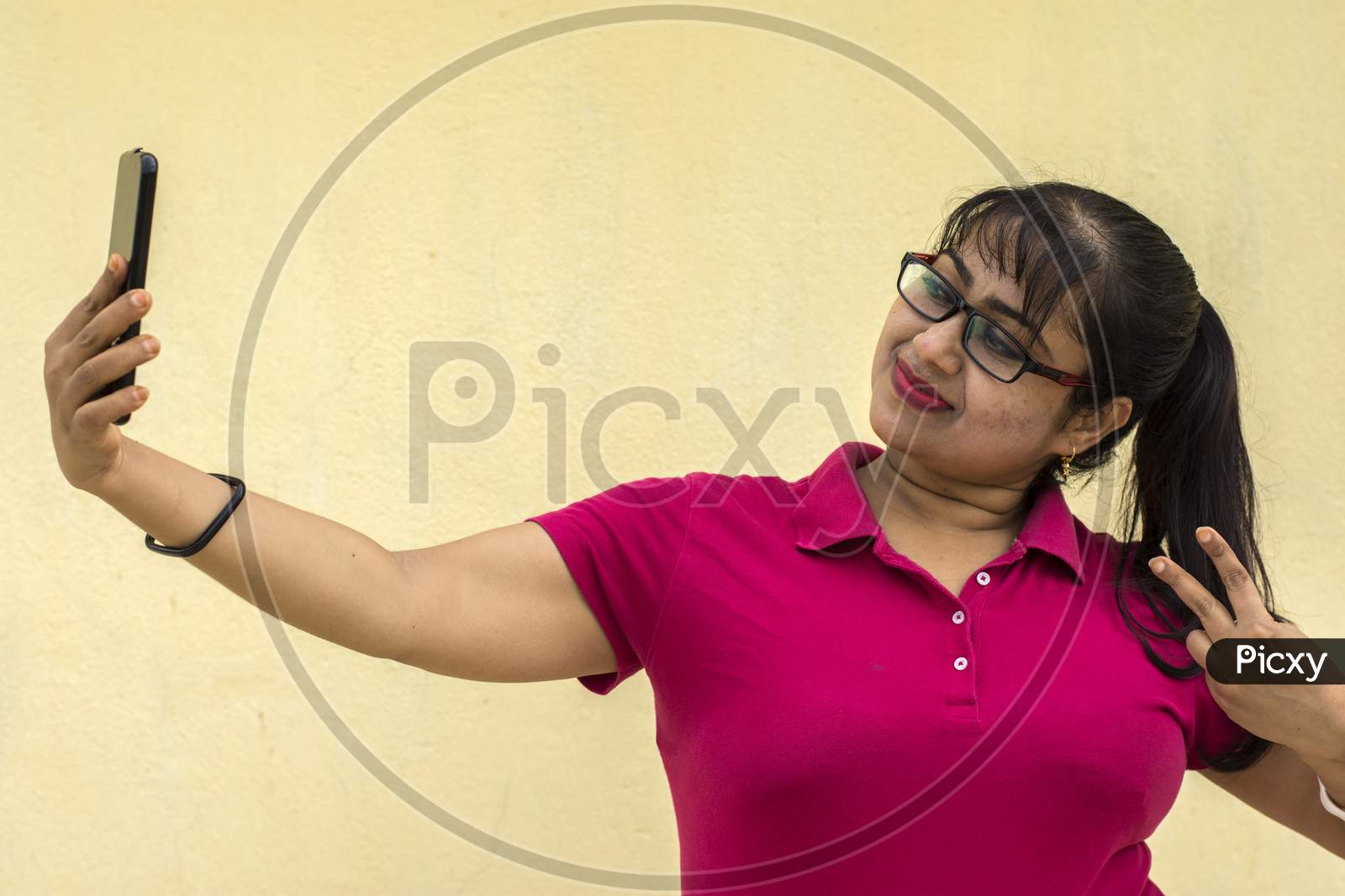 Indian Female Model Looking At Mobile Phone Taking Selfie Happily With Slight Smiling Face In Yellow Background With Copy Space For Text