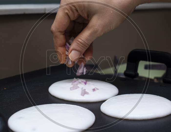 Putting Sliced Onions On Mini Round White Utappam With Fingers On Hot Non Stick Pan