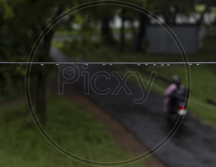 Selective Focus On The Water Droplets Hanging From The Cable In A Dark Wet Rainy Day While A Motor Cycle Passes By The Street